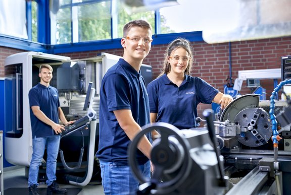Three young people stand in front of work machines in a workshop.